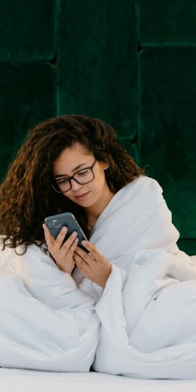Woman wrapped in white blanket using smartphone in cozy bedroom setting.